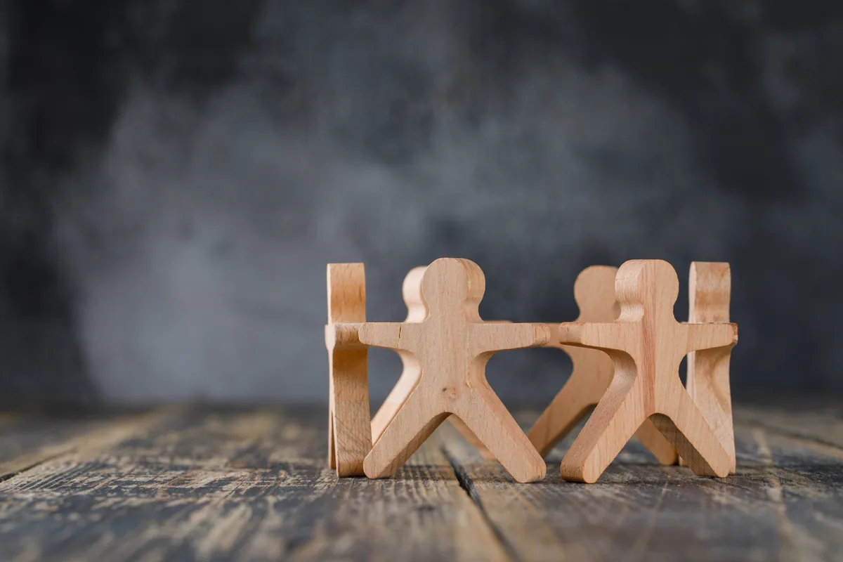 Holzfiguren stehen im Kreis und halten sich an den Händen, auf einer Holzfläche vor dunklem Hintergrund.