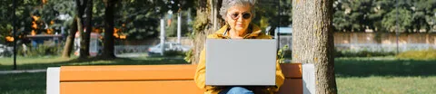 Eine ältere Frau in gelber Jacke sitzt mit einem aufgeklappten Laptop auf einer Parkbank.