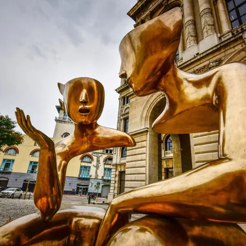 Zwei goldene Figuren einander zugewandt vor einem Altbau — die eine mit geöffnetem Mund, die andere mit erhobener Hand.